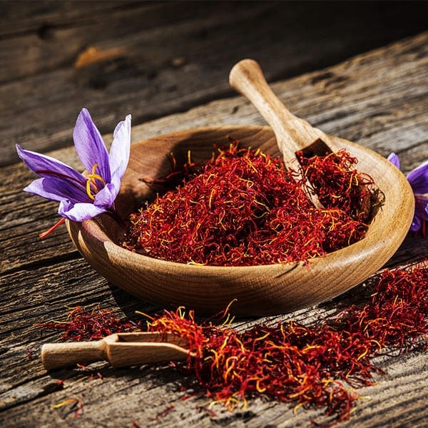 A wooden bowl filled with fresh saffron threads and a small wooden spoon, with purple saffron flowers scattered around.